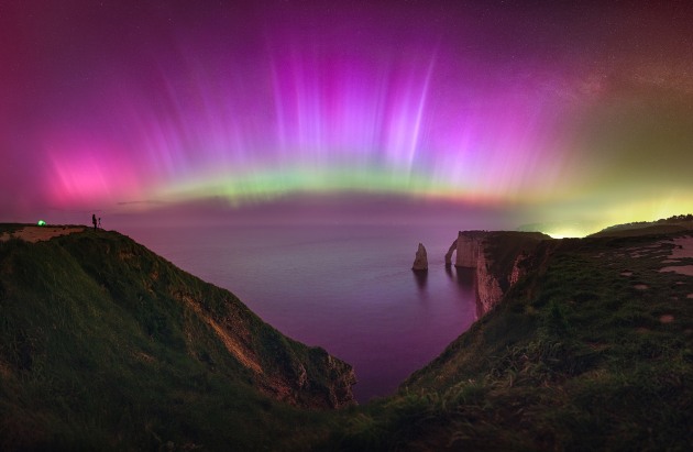 “A Historic Night: Northern Lights at Étretat” – Julien Looten, Normandy, France.
This rare G5 solar storm was observable across France at mid-latitudes, a region unaccustomed to such displays. For two years, I traveled regularly to the cliffs of Étretat (latitude 49.43), several hours from home, hoping to capture the aurora over this iconic landscape. After many unsuccessful attempts, my perseverance was finally rewarded.
This panoramic image captures a sky filled with vibrant auroras stretching 180 degrees from the southwest to the northeast. The display includes hues of pink, violet, and blue, with a green band near the horizon.